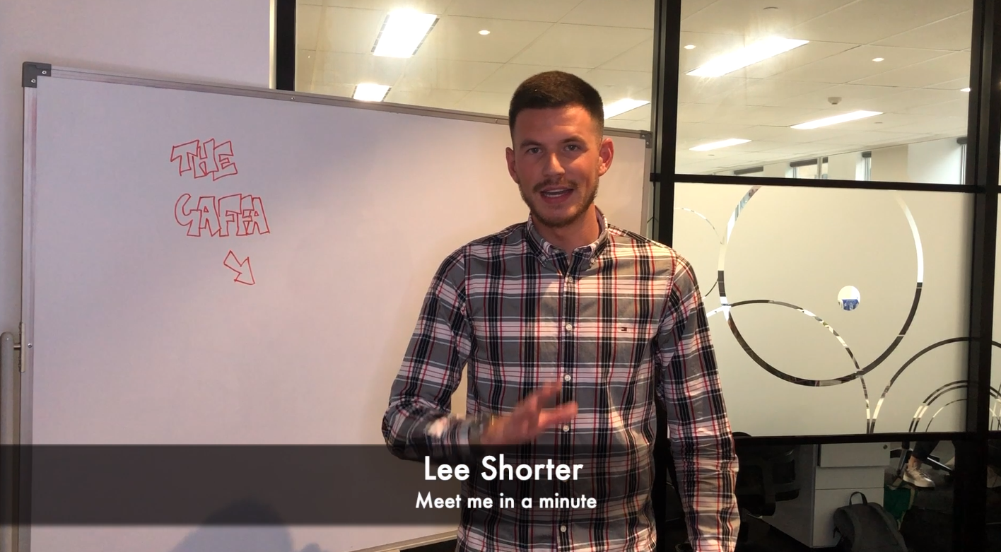 Man in a plaid shirt gesturing at the camera in an office, with a whiteboard to the left and glass partition behind. Overlay text reads 'Lee Shorter' and 'Meet me in a minute'.