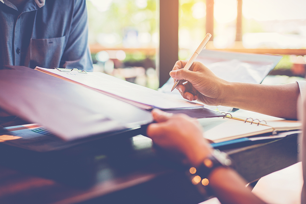 Two people at a desk review documents, one hand holding a pen ready to sign