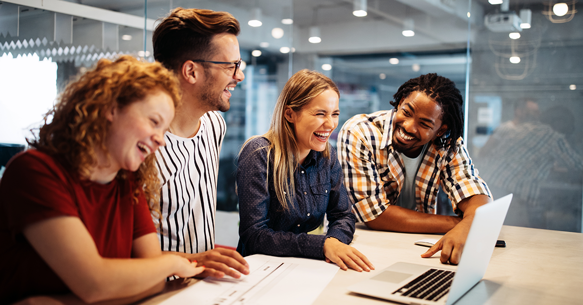 Four coworkers gathered around a laptop at a bright office, smiling during a collaboration.