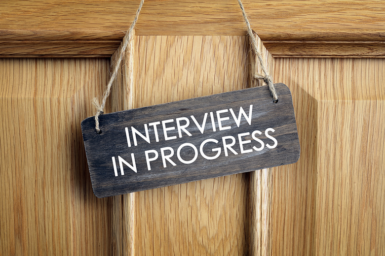 Dark wooden sign hanging on a door that reads 'INTERVIEW IN PROGRESS' indicating an ongoing interview session.
