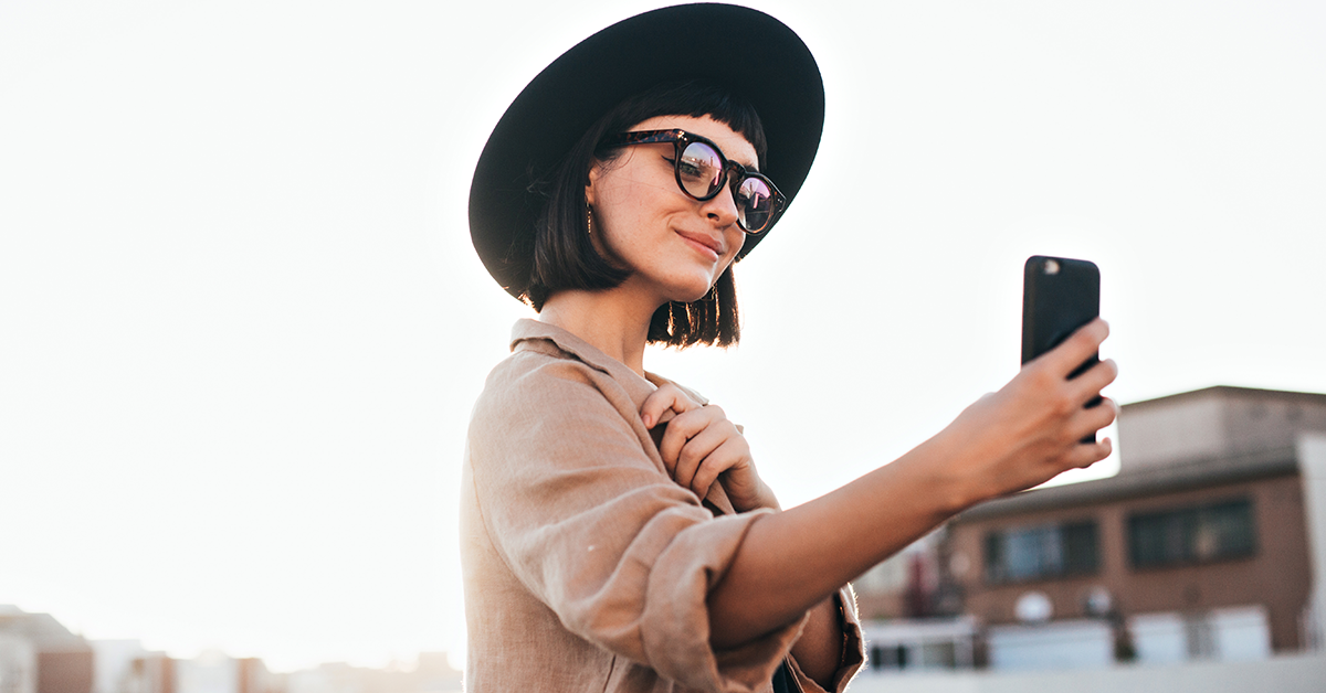 Woman wearing a wide-brimmed hat and sunglasses takes a selfie with her phone outdoors in an urban setting.